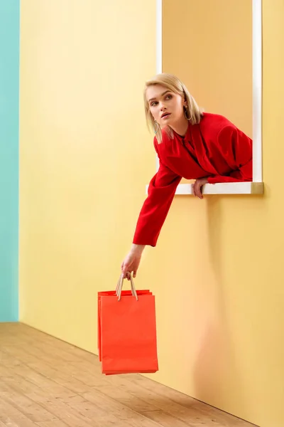 Mujer joven de moda en ropa roja con bolsas de compras rojas mirando por la ventana decorativa - foto de stock