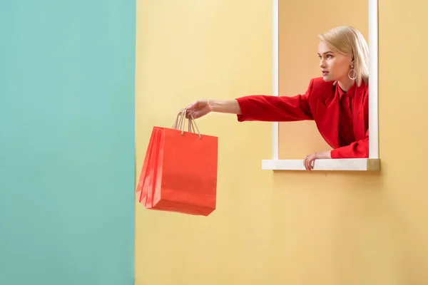 Young fashionable woman in red clothing with red shopping bags looking out decorative window — Stock Photo