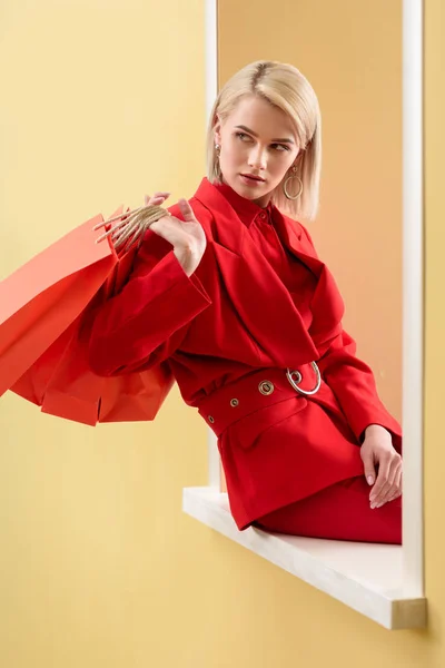 Young fashionable woman in red clothing with red shopping bags sitting on decorative window — Stock Photo