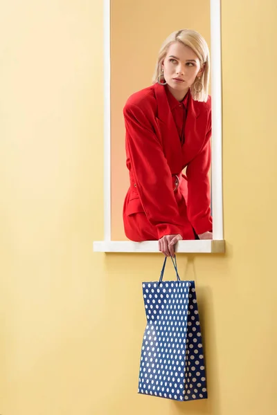 Stylish pensive woman in red suit with shopping bag with dots pattern in hand sitting on decorative window — Stock Photo