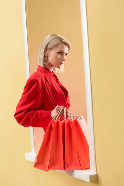 Jeune femme à la mode en vêtements rouges avec des sacs à provisions rouges assis sur la fenêtre décorative — Photo de stock