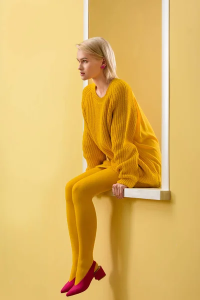 Вид сбоку стильной женщины в розовых туфлях, желтом свитере и колготках, сидящей на декоративном витрине — стоковое фото