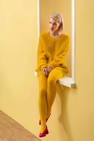 Stylish pensive woman in yellow sweater and tights sitting on decorative window — Stock Photo