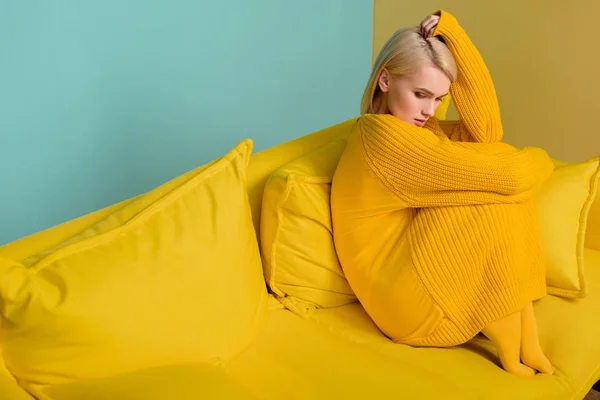 Side view of young blond woman in yellow sweater and tights sitting on yellow sofa on blue backdrop — Stock Photo