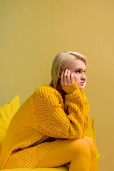 Vista lateral de la joven rubia en suéter amarillo y medias sentadas en un sofá amarillo - foto de stock