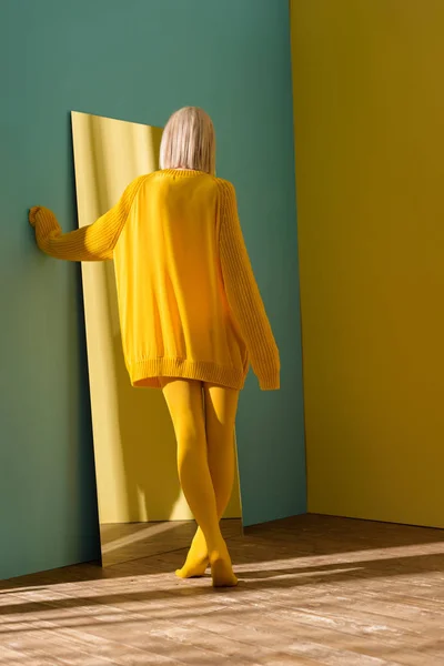 Задний вид блондинки в желтом свитере и колготках, стоящих перед зеркалом — стоковое фото