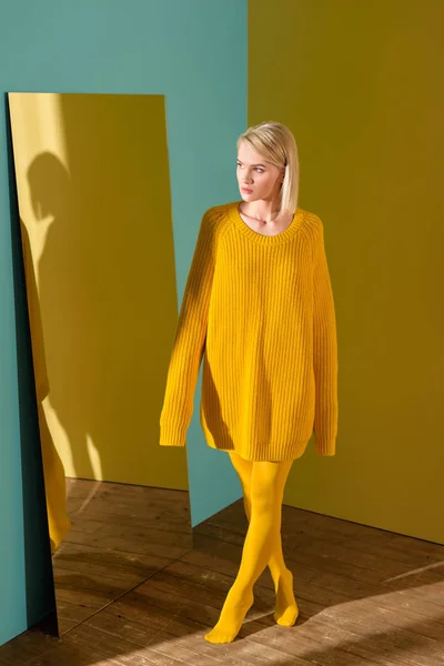 Hermosa mujer rubia pensativa en suéter amarillo y medias de pie en el espejo - foto de stock
