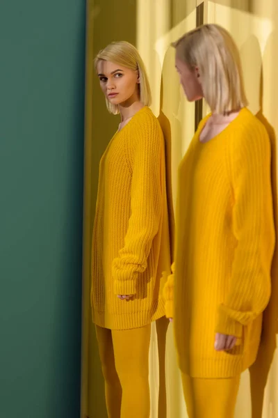 Blond woman in yellow sweater and tights looking at her reflection in mirror — Stock Photo