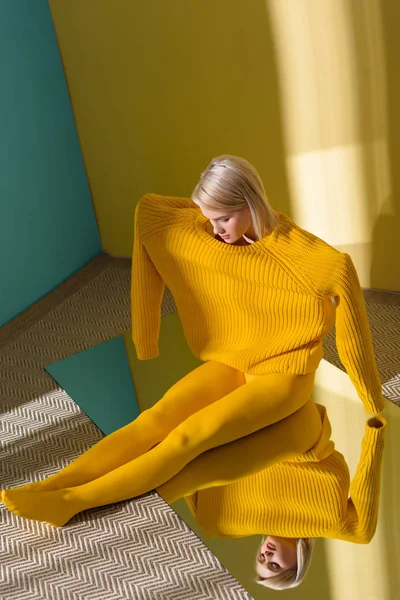 High angle view of attractive woman in yellow sweater and tights sitting on mirror with reflection in it — Stock Photo