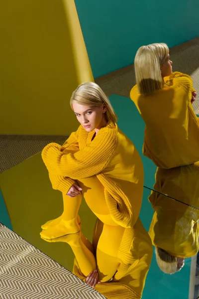 Hochwinkelaufnahme einer attraktiven nachdenklichen Frau in gelbem Pullover und Strumpfhose, die auf einem Spiegel sitzt und darin reflektiert — Stockfoto