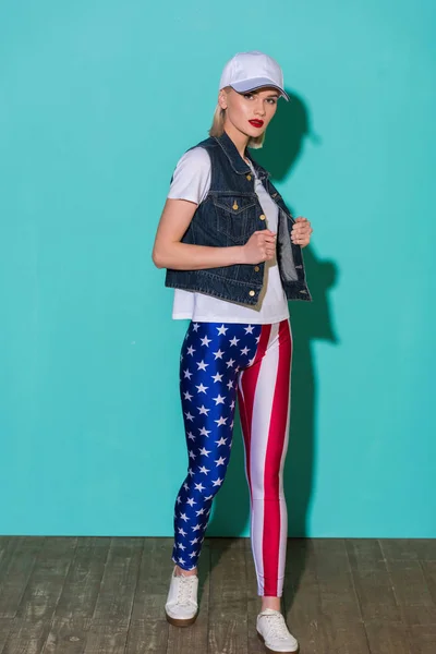 Stylish young woman in cap, white shirt, denim jacket and leggings with american flag pattern posing on blue background — Stock Photo