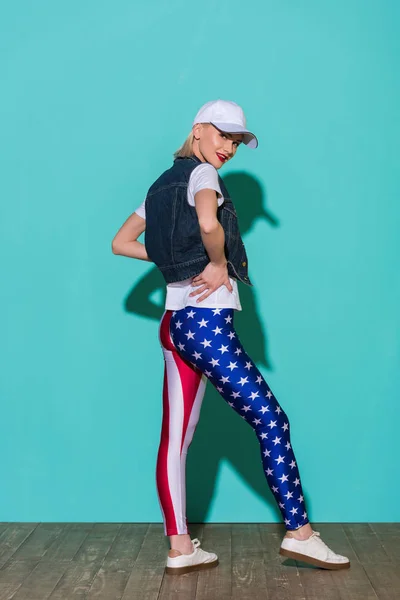 Jovem elegante em cap, camisa branca, jaqueta de ganga e leggings com padrão de bandeira americana posando no fundo azul — Fotografia de Stock