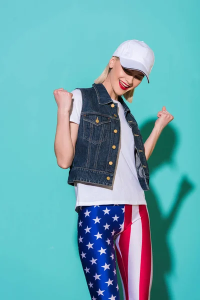 Cheerful stylish woman in cap, white shirt, denim jacket and leggings with american flag pattern posing on blue background — Stock Photo