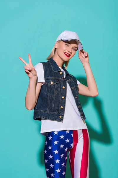 Stylish smiling woman in cap, white shirt, denim jacket and leggings with american flag pattern showing peace sign on blue background — Stock Photo