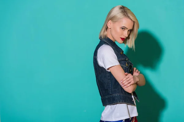 Vista lateral de la mujer joven con estilo en camisa blanca y chaqueta de mezclilla posando sobre fondo azul - foto de stock