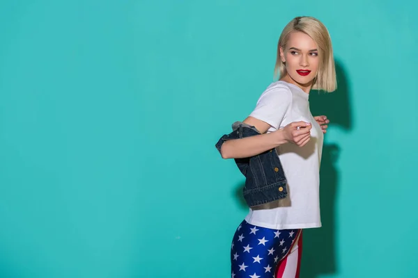 Vue latérale de la jeune femme élégante en chemise blanche, veste en denim et leggings avec motif drapeau américain sur fond bleu — Photo de stock