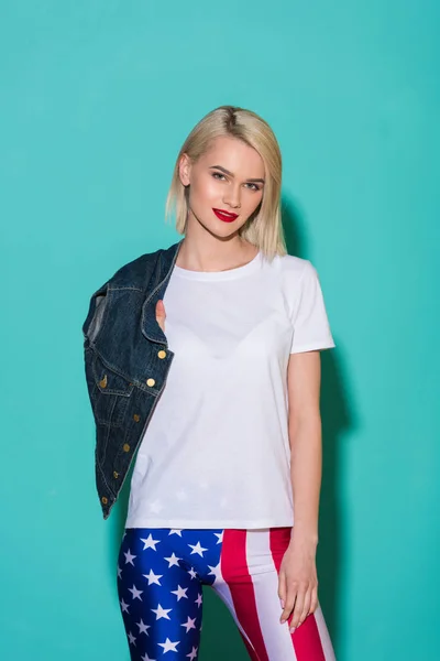 Portrait of stylish blond woman in white shirt and legging with american flag pattern with denim jacket posing on blue backdrop — Stock Photo