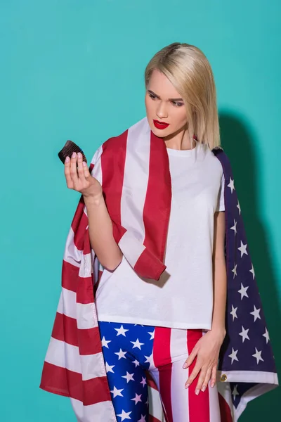 Portrait of young woman with american flag and cupcake on blue backdrop, celebrating 4th july concept — Stock Photo