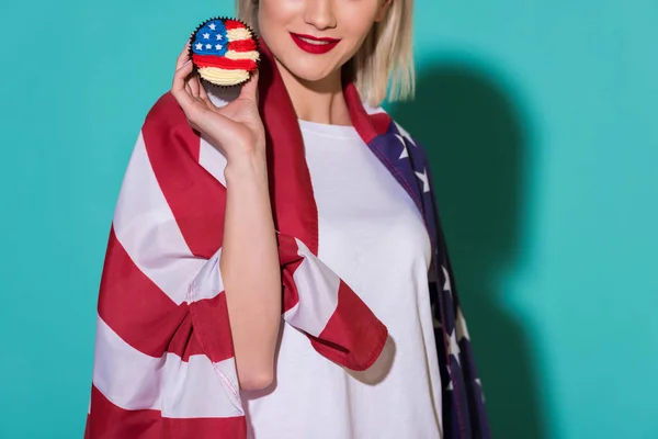 Tiro recortado de mulher sorridente com bandeira americana e cupcake na mão em pano de fundo azul, celebrando o conceito de 4 de julho — Fotografia de Stock