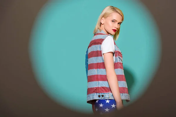 Side view of stylish young woman in denim jacket with american flag pattern posing on blue backdrop — Stock Photo