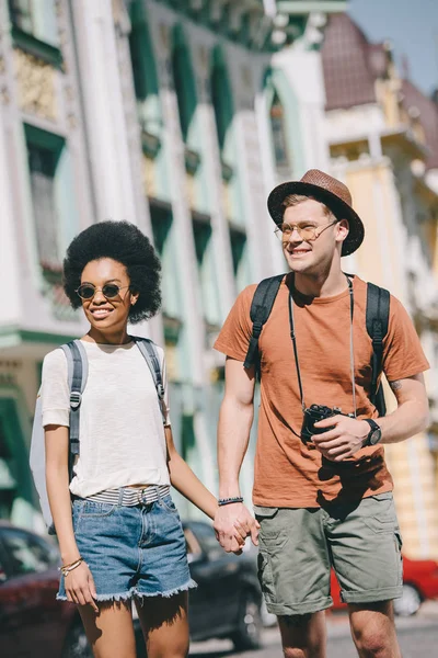 Sorrindo casal multicultural de viajantes com câmera e mochilas segurando as mãos uns dos outros — Fotografia de Stock