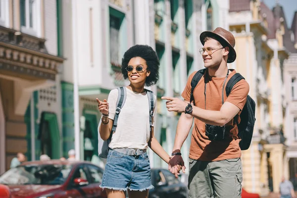 Smiling interracial couple of travelers pointing by fingers to each other — Stock Photo