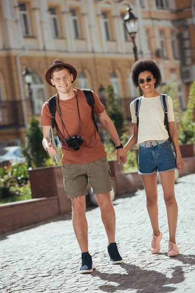 Pareja multicultural de turistas con cámara y mapa caminando y tomados de la mano el uno del otro - foto de stock