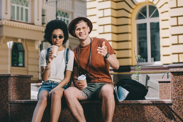 Male tourist with paper cup of coffee pointing to african american girlfriend — Stock Photo