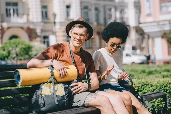 Multiethnic couple of young tourists with bottles of water resting on bench — Stock Photo