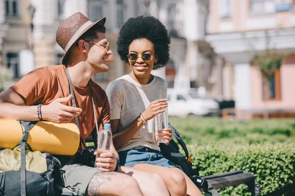 Happy interracial couple of tourists with bottles of water sitting on bench — Stock Photo