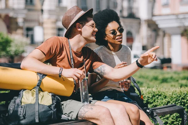 Junger männlicher Reisender mit Flasche Wasser zeigt mit dem Finger auf afrikanisch-amerikanische Freundin — Stockfoto