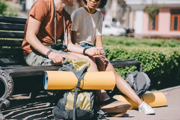 Cropped image of couple of travelers sitting on bench with backpacks and mats — Stock Photo