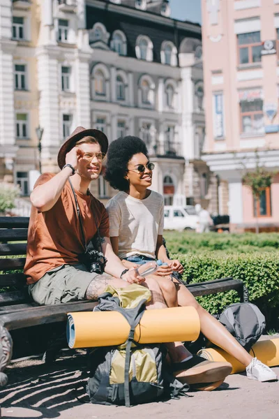 Coppia multiculturale di turisti con zaini e tappeti appoggiati su panca — Foto stock