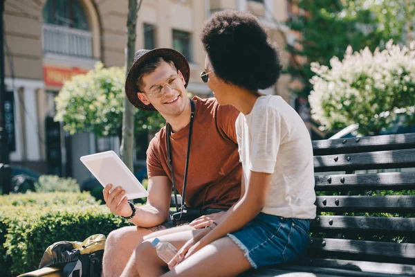 Pareja interracial de turistas sentados en el banco con la tableta digital - foto de stock