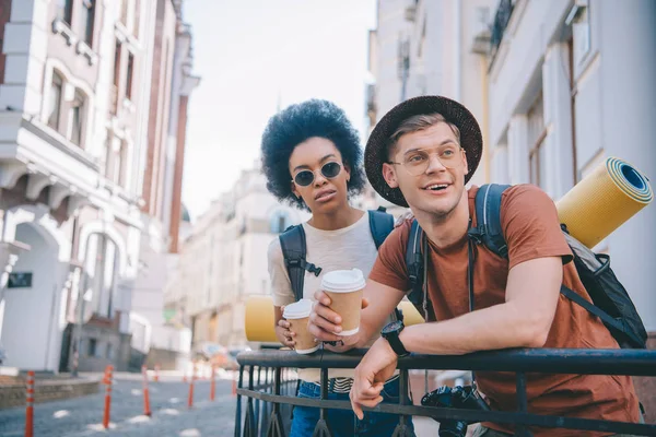 Pareja multicultural de turistas sosteniendo tazas de café y mirando hacia otro lado - foto de stock