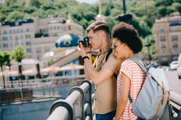 Vista laterale del viaggiatore maschile scattare foto sulla macchina fotografica e fidanzata afro-americana in piedi vicino — Foto stock