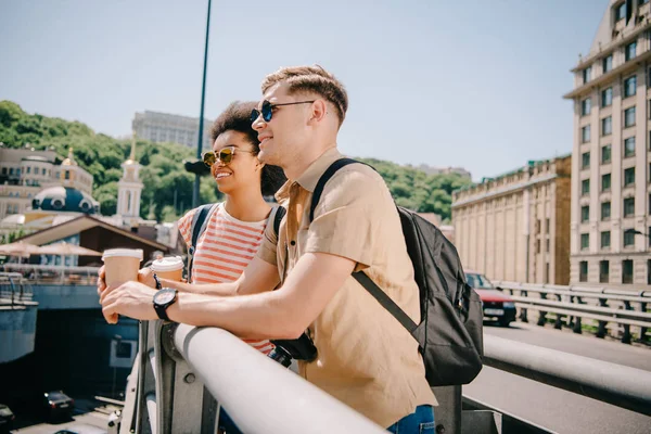 Multicultural happy couple of tourists with coffee cups looking at view from bridge — Stock Photo