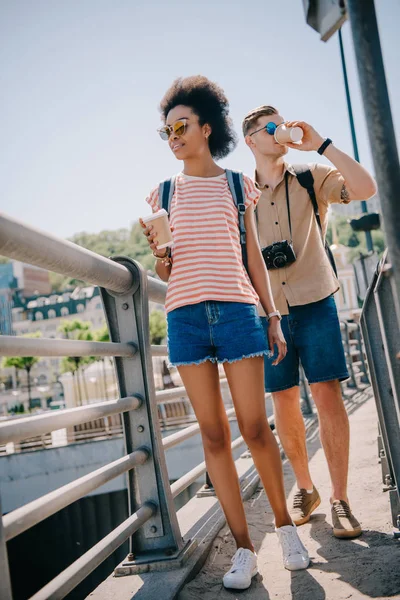 Interracial couple of tourists with disposable cups of coffee and camera walking on bridge — Stock Photo