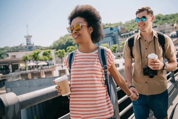 Pareja interracial de turistas con tazas de papel de café y cámara de mano y caminando en el puente - foto de stock