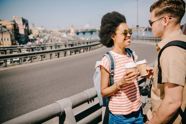 Sonriente afroamericana viajera y su novio con tazas de papel de café hablando entre sí - foto de stock