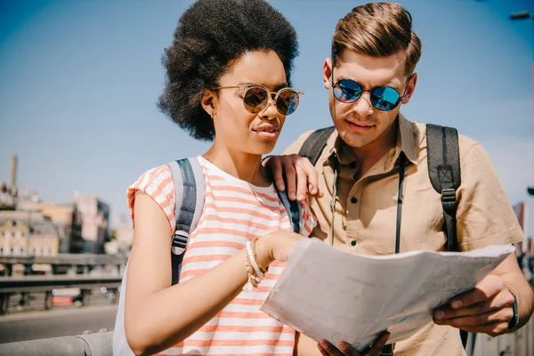 Мультикультурна пара туристів в сонцезахисних окулярах дивиться на мапу — стокове фото