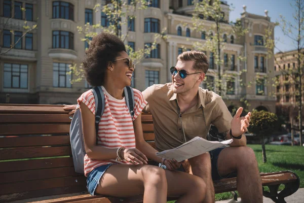 Sorridente coppia multietnica di turisti con mappa parlando tra loro in panchina — Foto stock