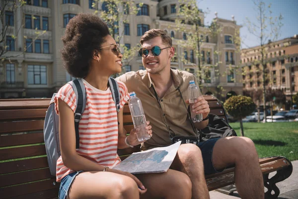 Coppia multiculturale di viaggiatori con mappa e bottiglia d'acqua appoggiata su panca — Foto stock