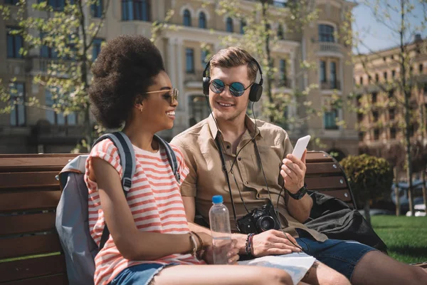 Stylish couple with camera, headphones and smartphone sitting on bench — Stock Photo