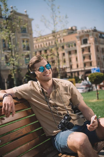 Hombre elegante con cámara escuchando música en auriculares y sosteniendo el teléfono inteligente en el banco - foto de stock