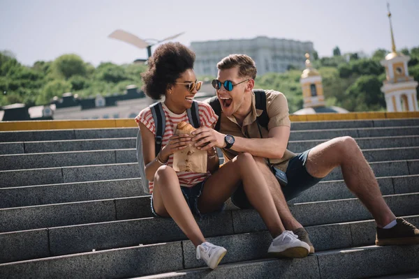 Young male traveler trying to eat girlfriend croissant on stairs — Stock Photo