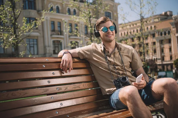 Joven en gafas de sol con cámara, auriculares y smartphone sentado en el banco - foto de stock