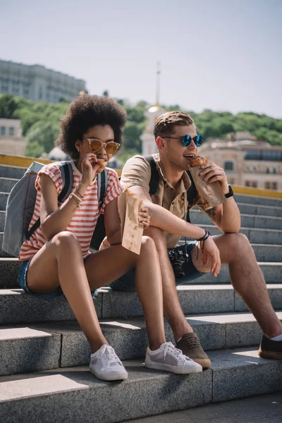 Multiethnic couple of travelers sitting on stairs and eating croissants — Stock Photo