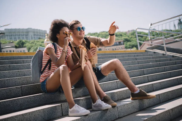 Jovem casal elegante em óculos de sol sentado nas escadas e comer croissants — Fotografia de Stock