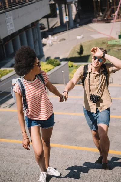 Junger männlicher Reisender mit Kamera und stylischer afrikanisch-amerikanischer Freundin hält Händchen und geht nach oben — Stockfoto